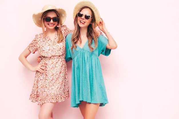 トレンディな夏のドレスを着た2人の若い美しい笑顔のブルネットのヒップスターの女性ピンクの壁の近くでポーズをとるセクシーな屈託のない女性帽子とサングラスで陽気で幸せな楽しいポジティブモデル