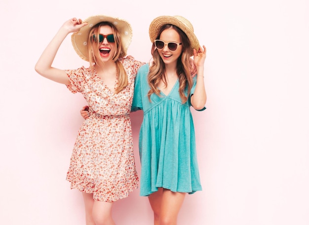 트렌디한 여름 드레스를 입은 두 젊은 아름다운 웃는 갈색 머리 힙스터 여성 분홍색 벽 근처에서 포즈를 취하는 섹시하고 평온한 여성 긍정적인 모델은 재미 있고 쾌활하고 행복합니다. 모자와 선글라스