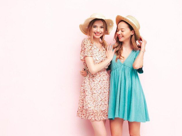Two young beautiful smiling brunette hipster female in trendy summer dresses Sexy carefree women posing near pink wall Positive models having fun Cheerful and happy In hats