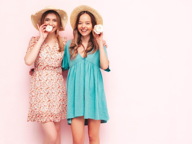 Two young beautiful smiling brunette hipster female in trendy summer dresses Sexy carefree women posing near pink wall Positive models having fun Cheerful and happy In hats