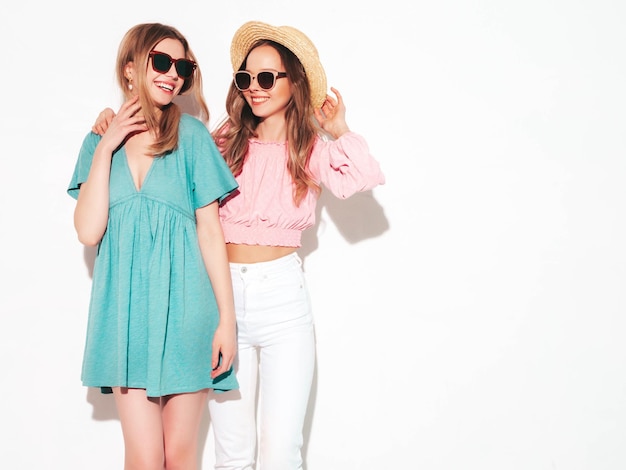 Two young beautiful smiling brunette hipster female in trendy summer dresses Sexy carefree women posing near white wall Positive models having fun Cheerful and happy In hats and sunglasses