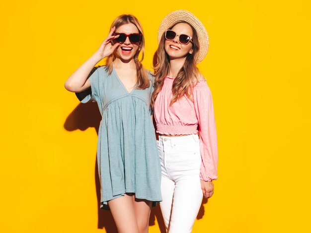 Two young beautiful smiling brunette hipster female in trendy summer dresses Sexy carefree women posing near yellow wall Positive models having fun Cheerful and happy In hats and sunglasses