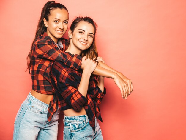 Two young beautiful smiling brunette hipster girls in trendy similar checkered shirt and jeans clothes.Sexy carefree women posing near pink wall in studio.Positive models having fun