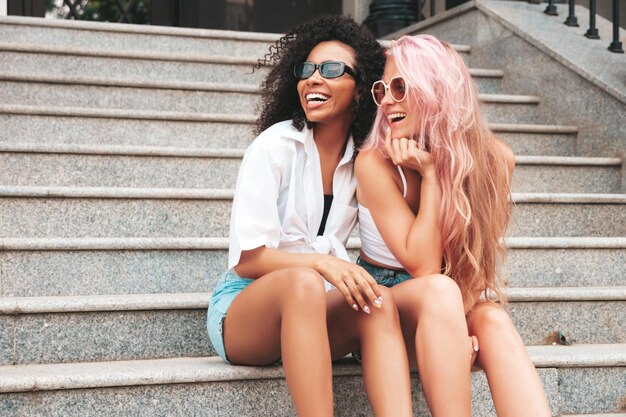 트렌디한 여름 옷을 입은 두 젊은 아름다운 미소 거리 배경에서 포즈를 취하는 섹시하고 평온한 다인종 여성선글라스를 쓴 긍정적인 모델 명랑하고 행복한 계단에 앉아