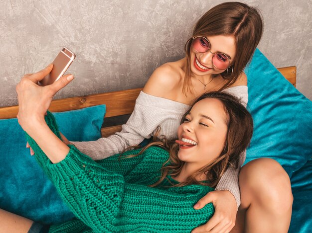 Two young beautiful smiling gorgeous girls in trendy summer clothes.  Sexy carefree women posing in interior and taking selfie. Positive models having fun with smartphone