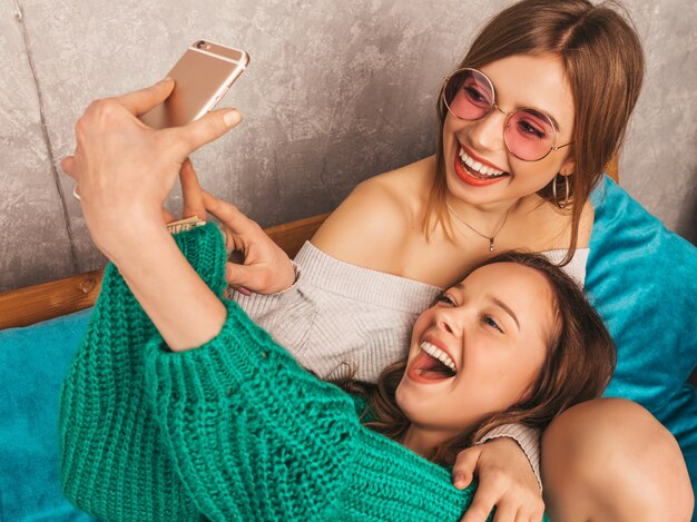 Two young beautiful smiling gorgeous girls in trendy summer clothes. Sexy carefree women posing in interior and taking selfie. Positive models having fun with smartphone