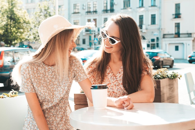 トレンディな夏の服を着た2人の若い美しい笑顔のヒップスターの女性ベランダカフェでおしゃべりし、お茶やコーヒーを飲むのんきな女性 Premium写真