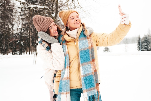 トレンディな暖かい服とスカーフで2人の若い美しい笑顔のヒップスターの女性。公園の路上でポーズをとる屈託のない女性。雪の中で楽しんでいるポジティブな純粋なモデル。冬のひとときを楽しんでいます。