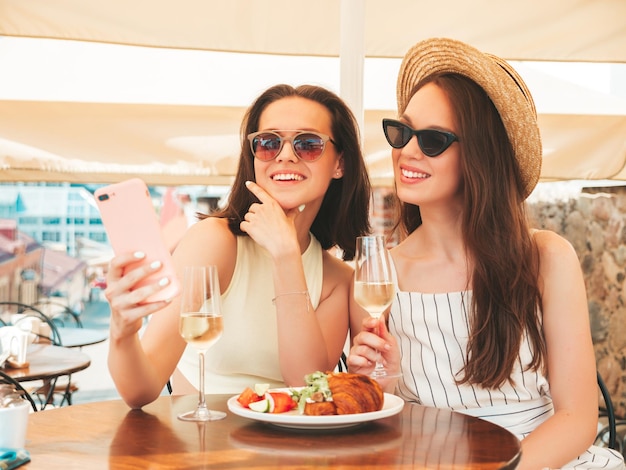 夏の服を着た2人の若い美しい笑顔のヒップスター女性通りのベランダカフェでポーズをとる屈託のない女性帽子で白ワインを飲むポジティブモデルアプリを使用してスマートフォンの画面を見る