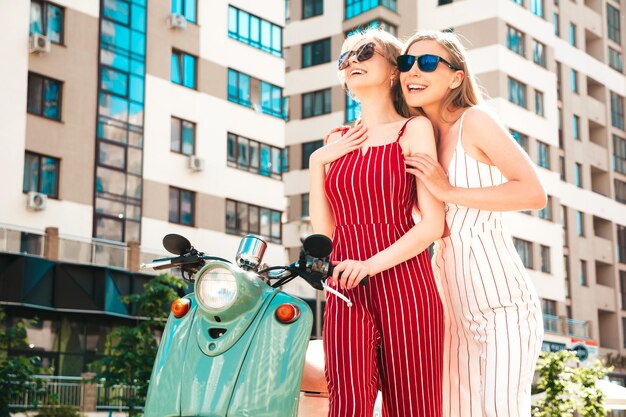 Two young beautiful smiling hipster female in trendy overallsSexy carefree women driving retro motorbike on the street backgroundPositive models having fun riding classic Italian scooter in eyewear