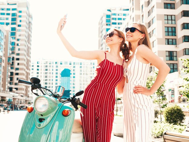 트렌디한 작업복을 입은 두 젊은 아름다운 힙스터 여성 거리 배경에서 복고풍 오토바이를 운전하는 섹시하고 평온한 여성 안경을 쓰고 고전적인 이탈리아 스쿠터를 타는 것을 즐기는 긍정적인 모델