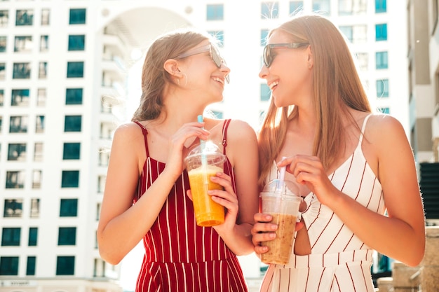 Two young beautiful smiling hipster female in trendy summer clothes Carefree women posing outdoorsPositive models holding and drinking fresh cocktail smoothie drink in plastic cup with straw