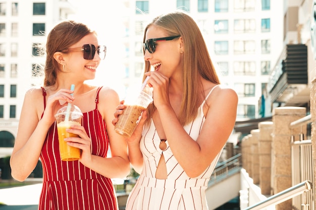 トレンディな夏の服を着た2人の若い美しい笑顔のヒップスター女性屋外でポーズをとるのんきな女性ストローとプラスチックカップで新鮮なカクテルスムージードリンクを保持し、飲むポジティブモデル