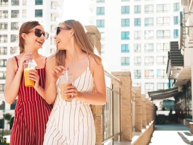 トレンディな夏の服を着た2人の若い美しい笑顔のヒップスター女性屋外でポーズをとるのんきな女性ストローとプラスチックカップで新鮮なカクテルスムージードリンクを保持し、飲むポジティブモデル