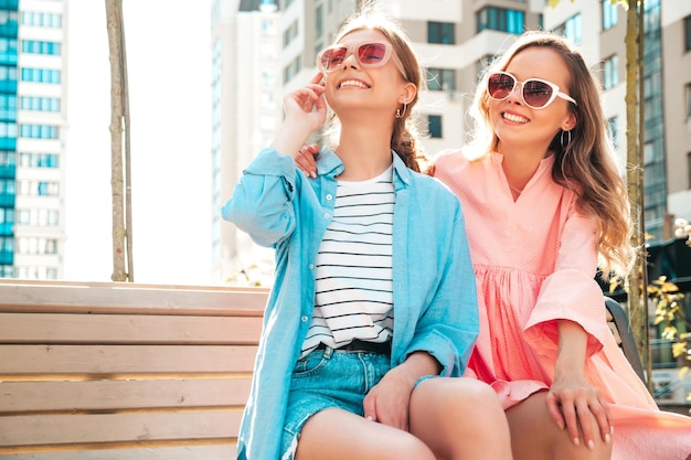 Две молодые красивые улыбающиеся хипстерши в модной летней одежде и платьеСексуальные беззаботные женщины позируют на улице Позитивные чистые модели веселятся на закате, сидя на скамейке в солнцезащитных очках