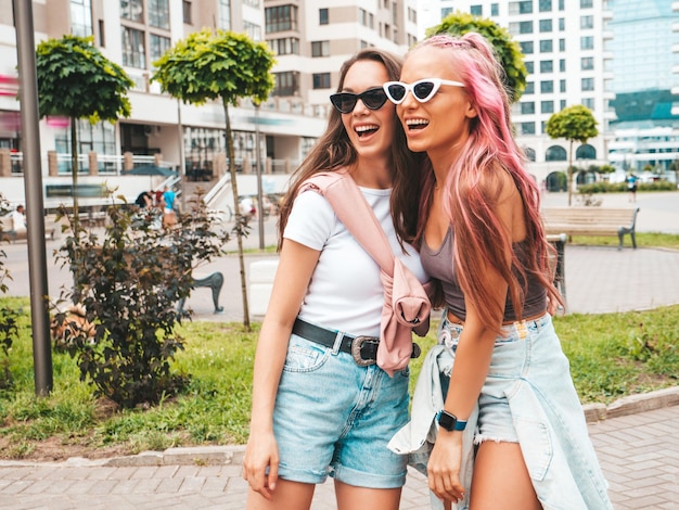 트렌디한 여름 옷을 입은 두 명의 젊고 아름다운 힙스터 여성. 분홍색 머리를 한 거리에서 포즈를 취한 평온한 섹시한 여성. 일몰을 즐기는 긍정적인 순수 모델. 명랑하고 행복한