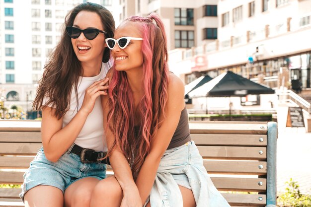 트렌디한 여름 옷을 입은 두 명의 젊고 아름다운 힙스터 여성. 분홍색 머리를 한 거리에서 포즈를 취한 평온한 섹시한 여성. 일몰을 즐기는 긍정적인 순수 모델. 명랑하고 행복한