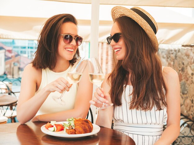 Две молодые красивые улыбающиеся женщины-хипстеры в модной летней одеждеБеззаботные женщины позируют в кафе на веранде на улицеПозитивные модели пьют белое вино в шляпеНаслаждаясь отпуском