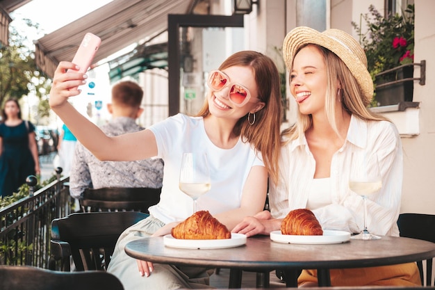 Two young beautiful smiling hipster female in trendy summer clothesCarefree women posing at veranda cafe in the streetPositive models drinking white wineEating croissantTaking selfie photos