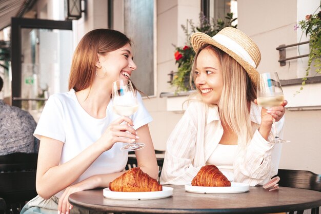 トレンディな夏服を着た2人の若い美しい笑顔のヒップスター女性通りのベランダカフェでポーズをとる屈託のない女性白ワインを飲むポジティブモデル休暇を楽しむクロワッサンを食べる