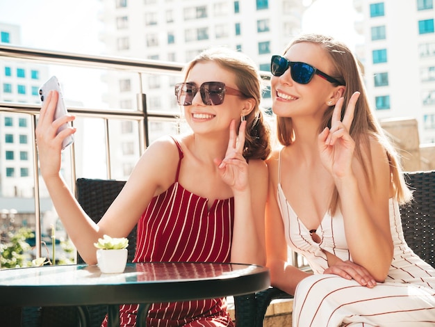 Two young beautiful smiling hipster female in trendy summer clothesCarefree women posing at veranda cafe in the streetPositive models having fun and taking selfie at smartphone