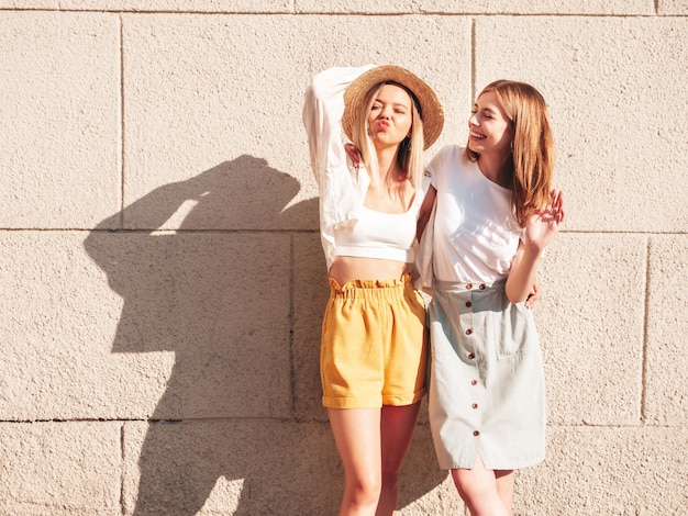 Две молодые красивые улыбающиеся хипстерши в модной летней одеждеСексуальные беззаботные женщины позируют возле белой стены на улице Позитивные чистые модели веселятся на закате, обнимаются и сходят с ума
