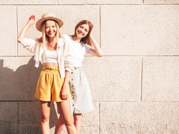 Две молодые красивые улыбающиеся хипстерши в модной летней одеждеСексуальные беззаботные женщины позируют возле белой стены на улице Позитивные чистые модели веселятся на закате, обнимаются и сходят с ума