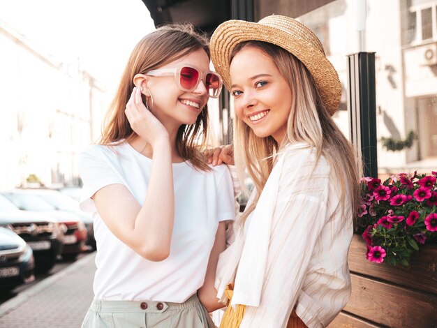 Two young beautiful smiling hipster female in trendy summer clothesSexy carefree women posing on the street background in hat Positive pure models having fun at sunset hugging