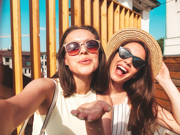Two young beautiful smiling hipster female in trendy summer clothesSexy carefree women posing on the street background in hat Positive pure models having fun at sunset taking Pov selfie