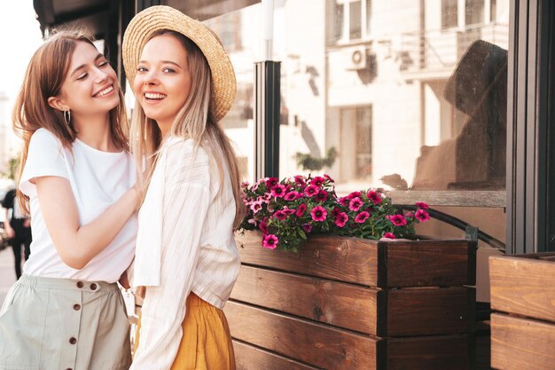 Две молодые красивые улыбающиеся хипстерши в модной летней одеждеСексуальные беззаботные женщины позируют на улице в шляпе Позитивные чистые модели веселятся на закате