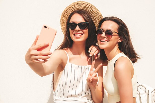 Two young beautiful smiling hipster female in trendy summer clothesSexy carefree women posing in the street near white wall in hat Positive pure models having fun at sunset taking selfie