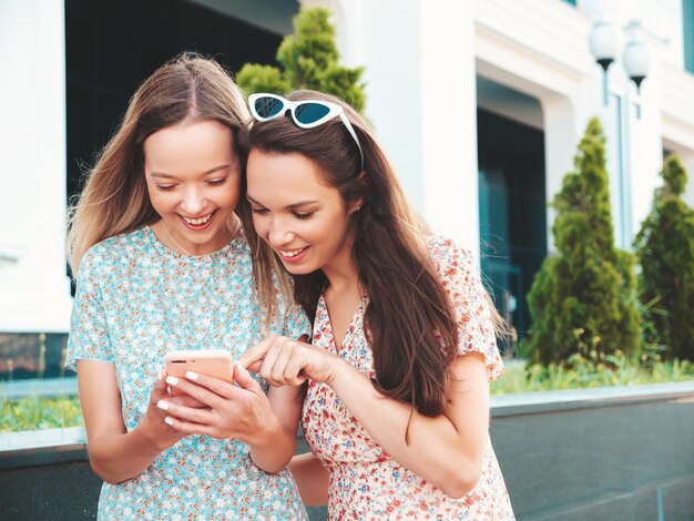 トレンディな夏服を着た2人の若い美しい笑顔のヒップスター女性通りでポーズをとるセクシーな屈託のない女性ハグを楽しんでいるポジティブモデル彼らは電話アプリを使用してスマートフォンの画面を見ています