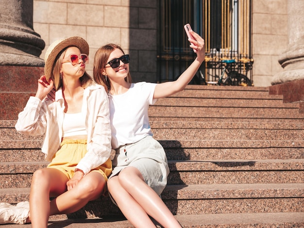 트렌디한 여름 옷을 입고 웃고 있는 두 젊은 힙스터 여성 거리에서 포즈를 취하는 섹시하고 평온한 여성 포옹을 즐기는 긍정적인 모델사진 셀카를 찍고 계단에 앉아
