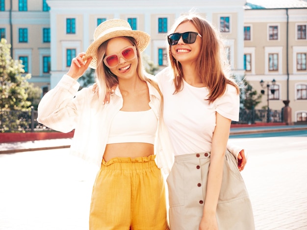 Due giovani belle donne hipster sorridenti in abiti estivi alla moda donne spensierate sexy che posano per strada modelli puri positivi che si divertono al tramonto con il cappello che si abbraccia e impazzisce