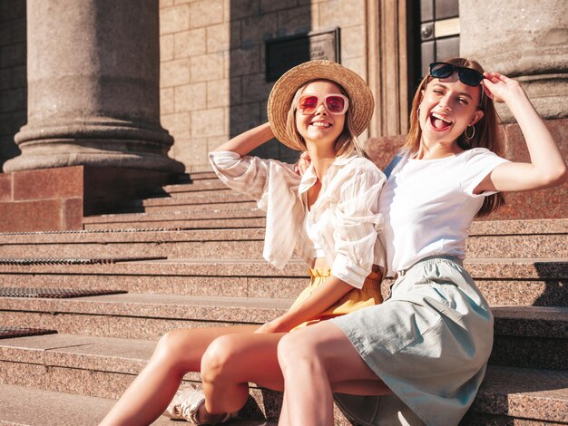 Две молодые красивые улыбающиеся хипстерши в модной летней одеждеСексуальные беззаботные женщины позируют на улице Позитивные чистые модели веселятся на закате, обнимаются и сходят с ума Сидя на лестнице