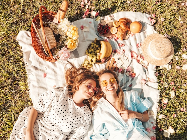 Two young beautiful smiling hipster female in trendy summer sundress and hats.Carefree women making picnic outside.