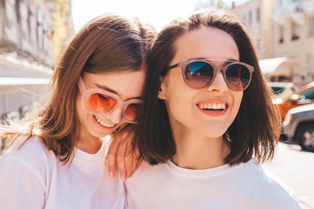 トレンディな夏の白いTシャツの服とジーンズで2人の若い美しい笑顔のヒップスターの女性。通りの背景でポーズをとるセクシーなのんきな女性。楽しんで、抱き締めて、夢中になっているポジティブなモデル