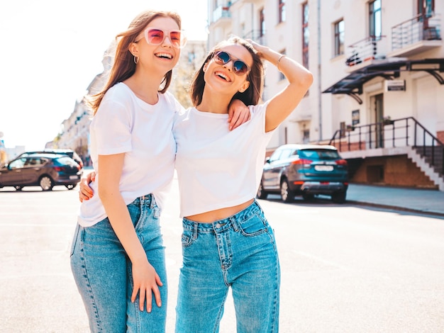 Две молодые красивые улыбающиеся женщины-хипстеры в модных летних белых футболках и джинсах. Сексуальные беззаботные женщины позируют на фоне улицы. Позитивные модели веселятся, обнимаются и сходят с ума