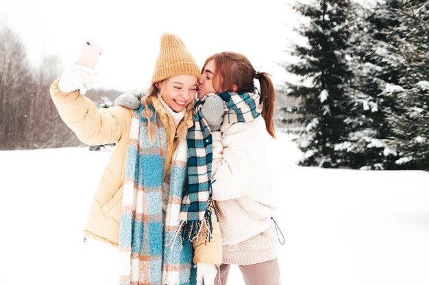 Two young beautiful smiling hipster female in trendy warm clothes and scarfs.Carefree women posing in the street in park. Positive pure models having fun in snow. Enjoying winter moments.Taking selfie