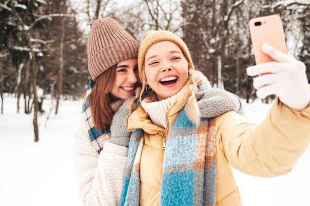 Two young beautiful smiling hipster female in trendy warm clothes and scarfs.Carefree women posing in the street in park. Positive pure models having fun in snow. Enjoying winter moments.Taking selfie