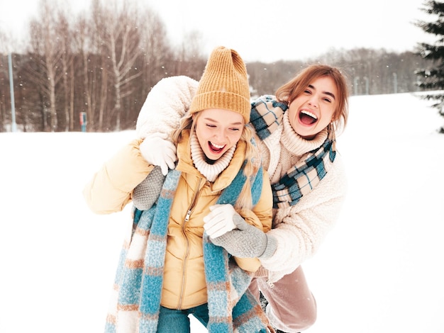 Two young beautiful smiling hipster female in trendy warm clothes and scarfs.Carefree women posing in the street in park. Positive pure models having fun in snow. Enjoying winter moments. Xmas concept