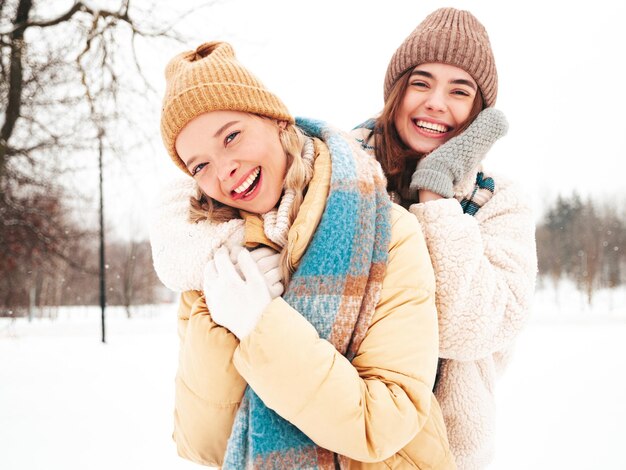 Two young beautiful smiling hipster female in trendy warm clothes and scarfs.Carefree women posing in the street in park. Positive pure models having fun in snow. Enjoying winter moments. Xmas concept