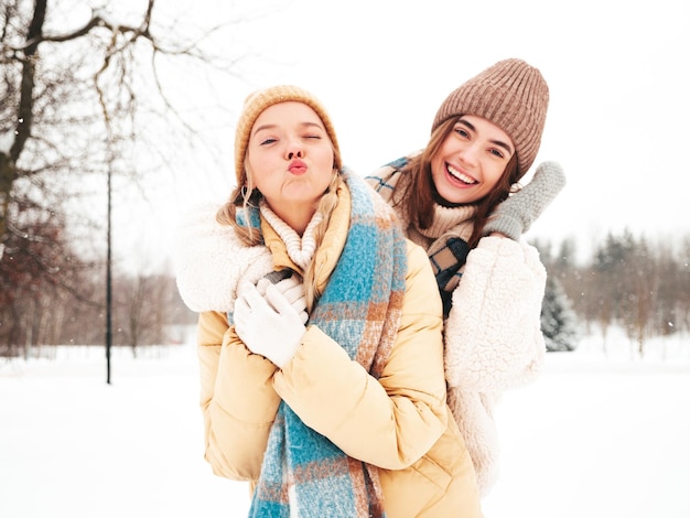 Two young beautiful smiling hipster female in trendy warm clothes and scarfs.Carefree women posing in the street in park. Positive pure models having fun in snow. Enjoying winter moments. Xmas concept