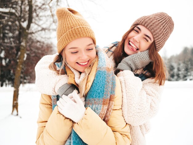 Две молодые красивые улыбающиеся хипстерские девушки в модной теплой одежде и шарфах. Беззаботные женщины позируют на улице в парке. Позитивные чистые модели веселятся в снегу. Наслаждаемся зимними моментами. Рождественское понятие