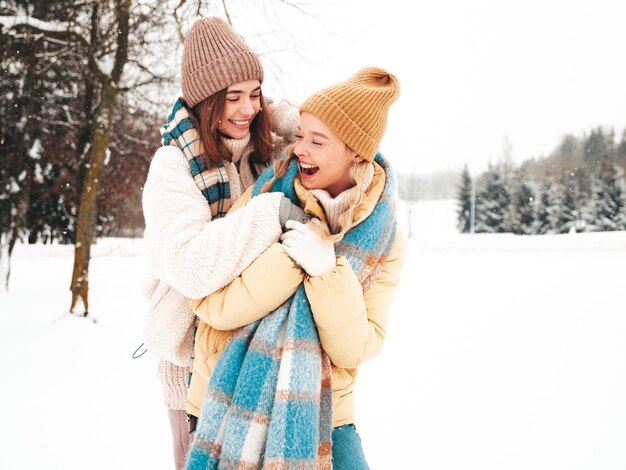 트렌디 한 따뜻한 옷과 스카프에 두 젊은 아름 다운 미소 hipster 여성. 공원에서 거리에서 포즈를 취하는 평온한 여성. 눈 속에서 즐겁게 노는 긍정적인 순수 모델. 겨울의 순간을 즐기고 있습니다. 크리스마스 컨셉