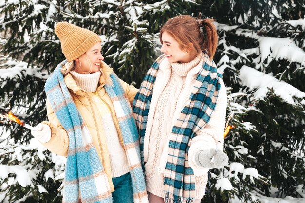 Two young beautiful smiling hipster female in trendy warm clothes and scarfs. carefree women posing in the street in park. positive pure models hugging each other. holding burning sparkling stick