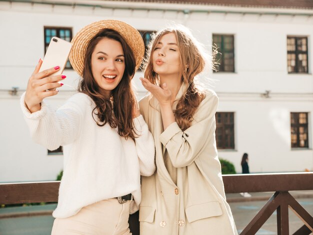 Две молодые красивые улыбающиеся хипстерские девушки в модном белом свитере и пальто