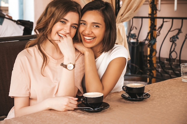 Two young beautiful smiling hipster girls in trendy summer casual clothes.Carefree women chatting in veranda terrace cafe and drinking coffee.Positive models having fun and communicating