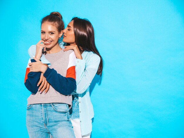 Two young beautiful smiling hipster girls in trendy summer casual clothes. Sexy women share secrets, gossip.Isolated on blue. Female bites her finger