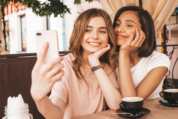 トレンディな夏服の2人の若い美しい笑顔の流行に敏感な女の子。ベランダカフェでおしゃべりとコーヒーを飲んで屈託のない女性。